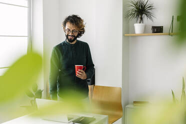 Glücklicher Freiberufler mit Kaffeetasse und Laptop am Schreibtisch im Büro - XLGF03149