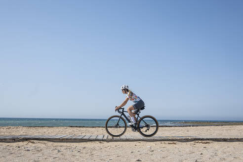 Cyclist riding bicycle at beach in front of blue sky on sunny day - JCMF02317