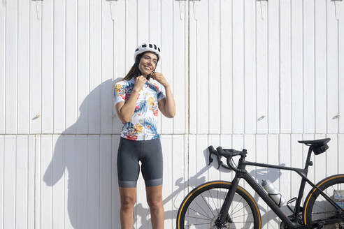 Glücklicher Radfahrer, der seinen Helm vor einer weißen Wand an einem sonnigen Tag einstellt - JCMF02319