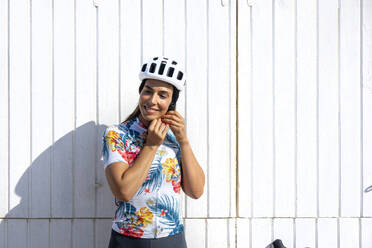 Glücklicher Radfahrer, der seinen Helm vor einer weißen Wand einstellt - JCMF02320