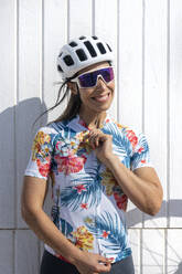 Glücklicher Radfahrer mit Sonnenbrille und Helm vor einer Mauer stehend - JCMF02325
