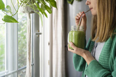 Frau trinkt grünen Smoothie am Fenster stehend zu Hause - SVKF00937
