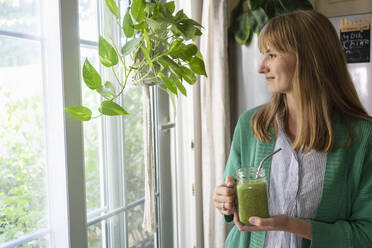 Lächelnde Frau mit grünem Smoothie, die zu Hause am Fenster steht - SVKF00938