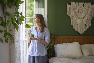 Thoughtful woman with green smoothie standing by window - SVKF00952