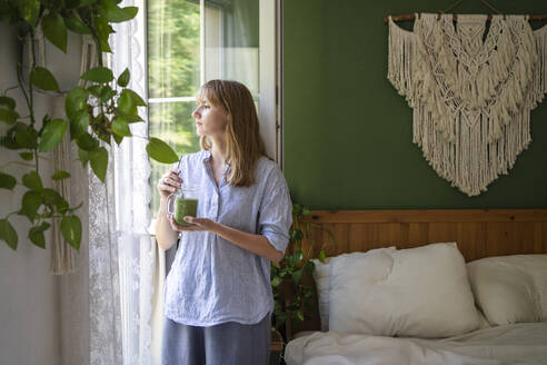 Nachdenkliche Frau mit grünem Smoothie am Fenster stehend - SVKF00952