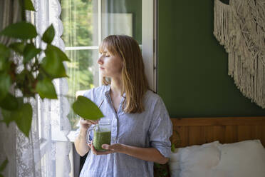 Woman with eyes closed holding smoothie standing by window in bedroom - SVKF00953