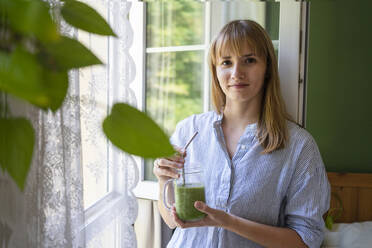 Lächelnde Frau mit Smoothie am Fenster stehend - SVKF00954