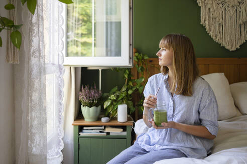 Nachdenkliche Frau mit Smoothie auf dem Bett sitzend zu Hause - SVKF00955