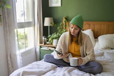 Woman using smart phone sitting with drink on bed at home - SVKF00959