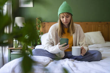 Sad woman using smart phone on bed with cup at home - SVKF00961