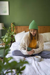 Woman wearing knit hat sitting on bed at home - SVKF00963