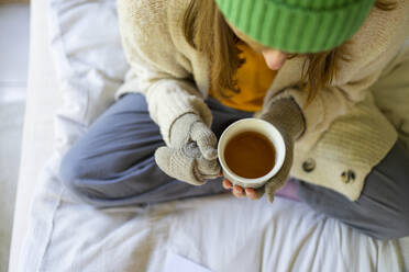 Woman wearing gloves sitting with tea cup on bed at home - SVKF00966