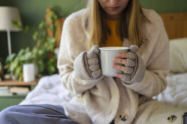 Woman wearing gloves holding tea cup on bed at home - SVKF00967