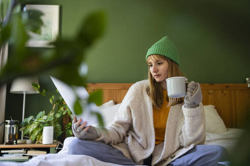 Woman examining financial bills sitting on bed at home - SVKF00971