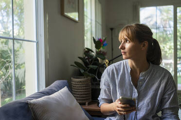 Woman with smart phone looking through window at home - SVKF00982