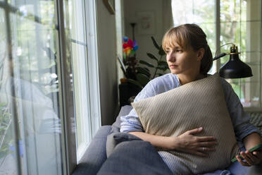 Nachdenkliche Frau mit Kissen, die zu Hause auf dem Sofa sitzt - SVKF00984