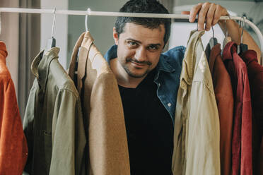 Smiling man standing behind clothes rack - VSNF00237