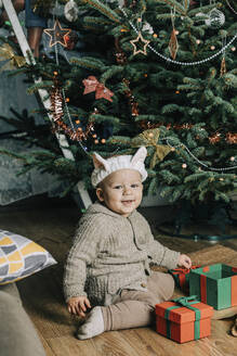 Glücklicher kleiner Junge mit Geschenkboxen vor einem Weihnachtsbaum sitzend - VSNF00243