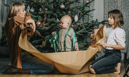 Mutter und Tochter halten Geschenkpapier mit einem kleinen Jungen, der vor einem Weihnachtsbaum steht - VSNF00246