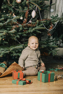 Lächelnder kleiner Junge mit Geschenkboxen vor einem Weihnachtsbaum sitzend - VSNF00253
