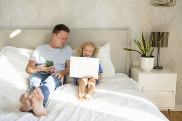 Smiling man with mobile phone looking at daughter using laptop on bed - SVKF00990