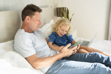 Father sharing mobile phone with daughter on bed in bedroom - SVKF00992