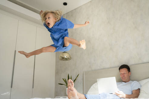 Playful girl jumping on bed in front of father with laptop at home - SVKF01002