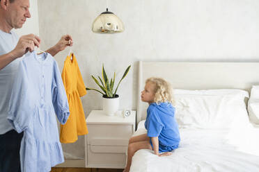 Father showing dresses to daughter sitting on bed at home - SVKF01003