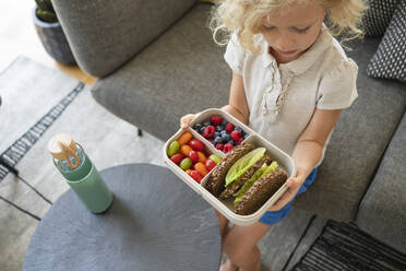 Girl holding lunch box with healthy food on sofa at home - SVKF01010