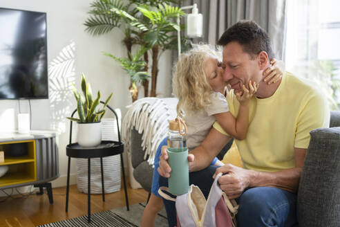 Mädchen küsst Vater mit Rucksack und Wasserflasche auf dem Sofa zu Hause - SVKF01013