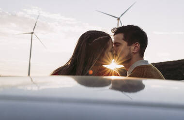 Couple kissing by car at sunset with wind farm in background - LJF02377