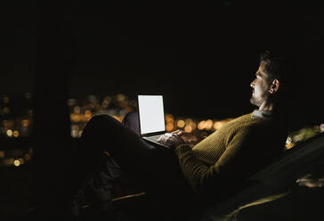 Mann sitzt nachts auf einem Hügel über einer beleuchteten Stadt und benutzt einen Laptop - LJF02382