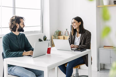 Happy business colleagues discussing with laptop at desk in workplace - XLGF03168