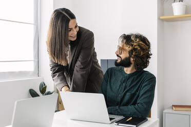 Smiling business colleagues looking at each other with laptop on desk in office - XLGF03173