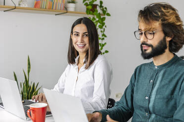 Smiling young businesswoman by colleague using laptop at desk - XLGF03187