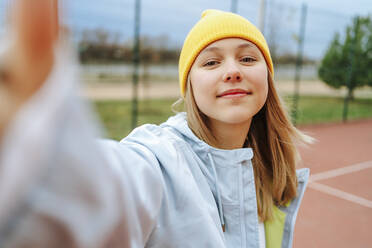 Lächelndes Mädchen mit Strickmütze macht ein Selfie auf dem Spielplatz - MDOF00436