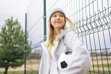 Smiling blond girl standing with soccer ball in front of fence - MDOF00441