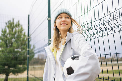 Lächelndes blondes Mädchen steht mit Fußball vor einem Zaun - MDOF00441