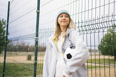 Contemplative girl wearing knit hat standing with soccer ball - MDOF00445