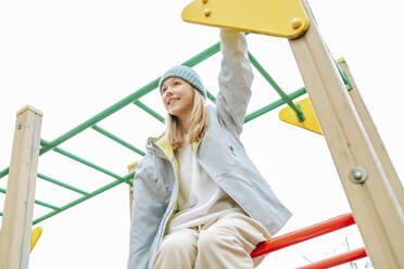 Glückliches Mädchen im Teenageralter sitzt auf dem Spielplatz auf einem Klettergerüst - MDOF00452