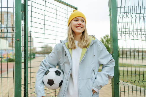 Glückliches Teenager-Mädchen mit gelber Strickmütze am Zaun eines Spielplatzes - MDOF00454