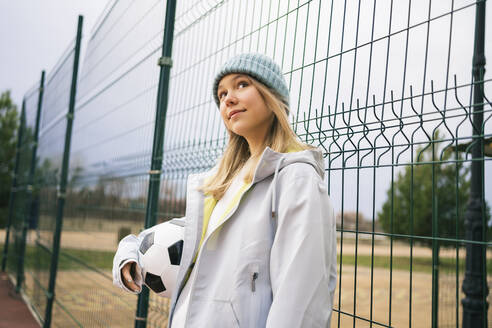 Nachdenkliches junges Mädchen mit Fußball vor einem Zaun stehend - MDOF00459