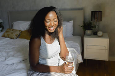Glückliche Frau mit geschlossenen Augen, die ein Trinkglas auf dem Bett zu Hause hält - TYF00515