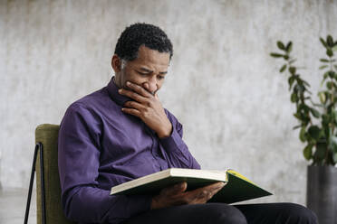 Mature man reading book on chair at home - EYAF02422