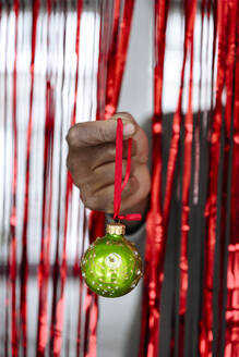 Hand eines Mannes, der eine grüne Weihnachtskugel vor einem roten Lametta-Vorhang hält - EYAF02462