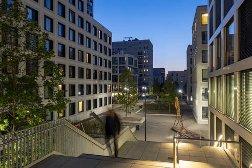 Deutschland, Bayern, München, Fußgänger, der in der Abenddämmerung eine Treppe hinaufgeht, mit Wohnungen und Bürogebäuden im Hintergrund - MAMF02354