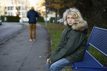 Blonder Junge sitzt auf einer Bank am Fußweg - NJAF00098