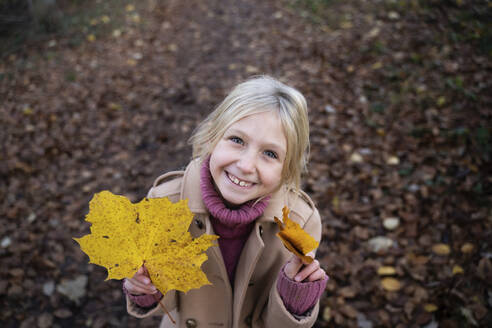 Glückliches blondes Mädchen stehend mit gelben Ahornblättern - NJAF00105