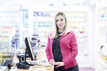 Apothekerin mit blondem Haar steht am Schreibtisch in einer Apotheke - NJAF00147