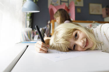 Lächelndes Mädchen, das einen Stift hält und sich auf ein Papier stützt, mit seiner Schwester im Hintergrund - TYF00530